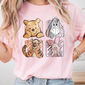 May include: A light pink t-shirt featuring four cartoon character portraits in square frames. The characters include a yellow bear, a gray donkey, an orange tiger, and a pink bunny. The shirt has short sleeves and a crew neck.