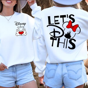 K&ouml;nnte beinhalten: Wei&szlig;es Sweatshirt mit dem Text "Disney vibes" und Mickey Mouse-H&auml;nden, die ein Herz bilden, auf der Vorderseite. Auf der R&uuml;ckseite steht der Text "Let's Do This" mit einer Minnie Mouse-Silhouette und roter Schleife. Ein l&auml;ssiges, themenbezogenes Kleidungsst&uuml;ck.