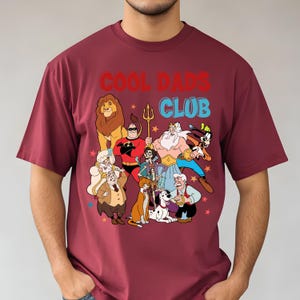 Pode incluir: Camiseta bord&ocirc; com o texto "COOL DADS CLUB" em vermelho e azul. A camiseta apresenta um gr&aacute;fico colorido de personagens de desenhos animados, incluindo um le&atilde;o, um super-her&oacute;i e v&aacute;rias outras figuras animadas. O design &eacute; contra um fundo de pequenas estrelas.