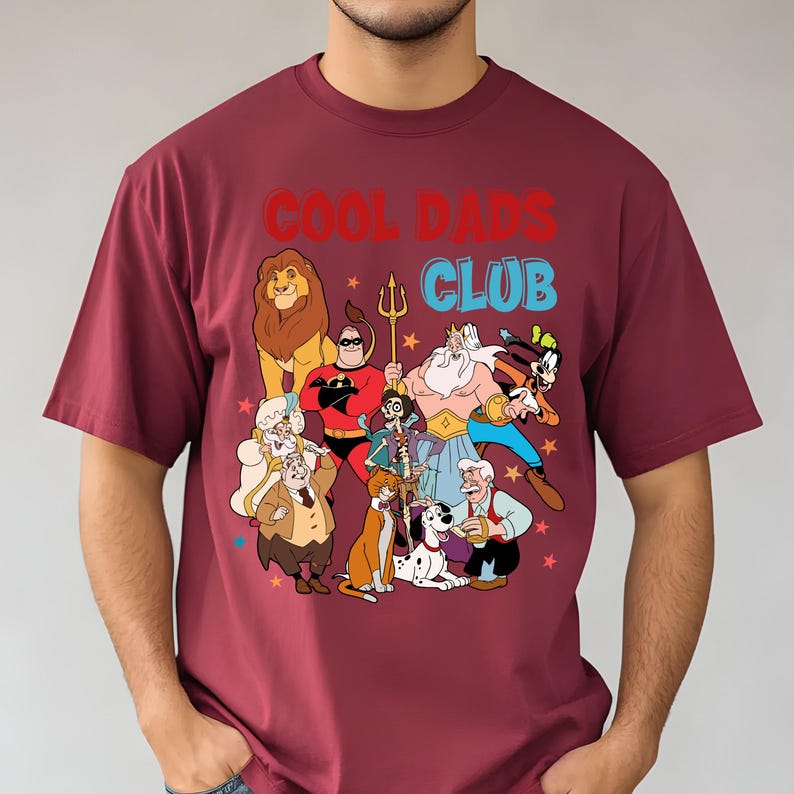 Pode incluir: Camiseta bord&ocirc; com o texto "COOL DADS CLUB" em vermelho e azul. A camiseta apresenta um gr&aacute;fico colorido de personagens de desenhos animados, incluindo um le&atilde;o, um super-her&oacute;i e v&aacute;rias outras figuras animadas. O design &eacute; contra um fundo de pequenas estrelas.