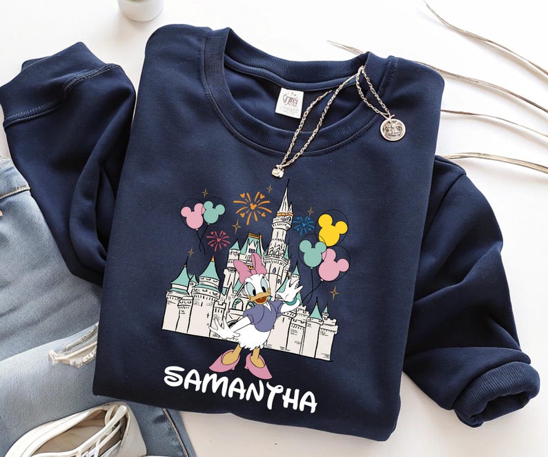 Może przedstawiać: Granatowa bluza z nadrukiem Kaczki Daisy przed zamkiem, fajerwerkami i balonami. Imię "SAMANTHA" jest wydrukowane poniżej. Srebrny naszyjnik jest ułożony na bluzie.