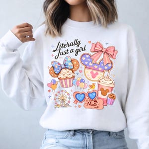 Pode incluir: Camisola branca com um design gr&aacute;fico colorido com um cupcake, orelhas do Mickey, uma roda gigante e o texto "Literally Just a girl". O design inclui cora&ccedil;&otilde;es, la&ccedil;os e um bilhete que diz "Let the Magic Begin!"