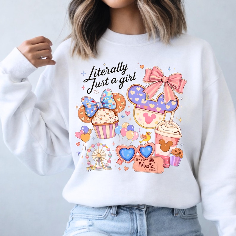 Pode incluir: Camisola branca com um design gr&aacute;fico colorido com um cupcake, orelhas do Mickey, uma roda gigante e o texto "Literally Just a girl". O design inclui cora&ccedil;&otilde;es, la&ccedil;os e um bilhete que diz "Let the Magic Begin!"