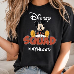Op de afbeelding: Zwart T-shirt met Mickey Mouse van Disney. Het ontwerp bevat het woord "Disney" in witte letters boven een afbeelding van Mickey Mouse. Daaronder staat het woord "SQUAD" in rood met een gele omlijning en de naam "KATHLEEN" in wit.