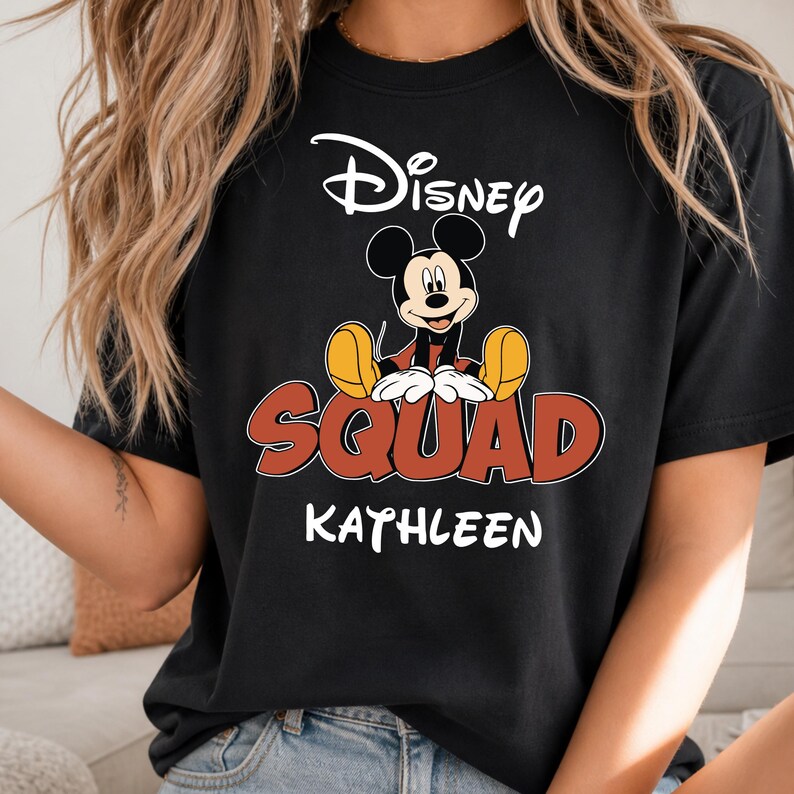 Op de afbeelding: Zwart T-shirt met Mickey Mouse van Disney. Het ontwerp bevat het woord "Disney" in witte letters boven een afbeelding van Mickey Mouse. Daaronder staat het woord "SQUAD" in rood met een gele omlijning en de naam "KATHLEEN" in wit.
