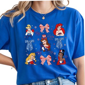 May include: A royal blue t-shirt with a patriotic design. It features cartoon princesses, bows, and fireworks in red, white, and blue. The design includes stars and bow ties, creating a festive look.