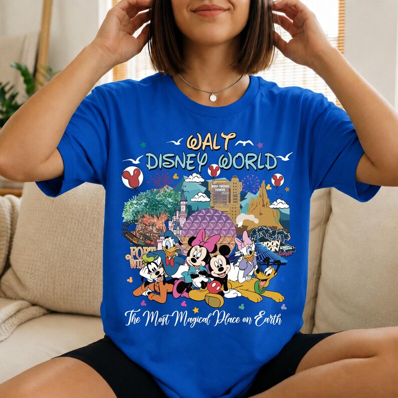 K&ouml;nnte beinhalten: K&ouml;nigsblaues T-Shirt mit einem farbenfrohen Aufdruck von Disney-Figuren und Wahrzeichen. Das Design zeigt Micky und Minnie Maus, Donald Duck und Goofy, mit dem Text "Walt Disney World" und "The Most Magical Place on Earth."