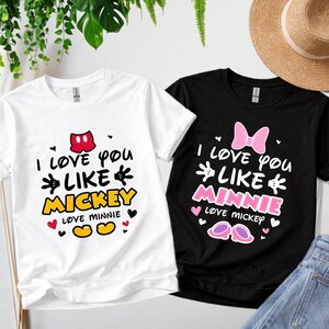 Pode incluir: Duas camisetas, uma branca e outra preta, com gr&aacute;ficos tem&aacute;ticos da Disney. A camiseta branca diz "I love you like Mickey" e a camiseta preta diz "I love you like Minnie". Ambas as camisetas t&ecirc;m cora&ccedil;&otilde;es e designs inspirados no Mickey Mouse.