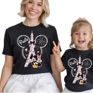 K&ouml;nnte beinhalten: Schwarze T-Shirts mit einem Design mit dem Eiffelturm, Mickey und Minnie Mouse und dem Wort "Paris". Das Design umfasst rosa Herzen und eine Schleife. Die T-Shirts werden von zwei Personen getragen.
