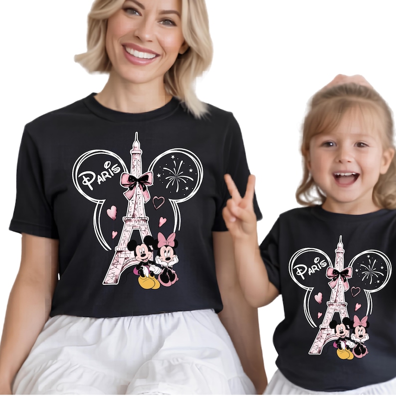 K&ouml;nnte beinhalten: Schwarze T-Shirts mit einem Design mit dem Eiffelturm, Mickey und Minnie Mouse und dem Wort "Paris". Das Design umfasst rosa Herzen und eine Schleife. Die T-Shirts werden von zwei Personen getragen.