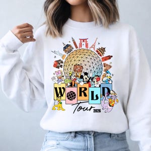 May include: White sweatshirt with a vibrant graphic of Disney characters and global landmarks. The design includes the text "WORLD Tour 2026", with Mickey Mouse, Donald Duck, and other characters. The design is set against a globe.