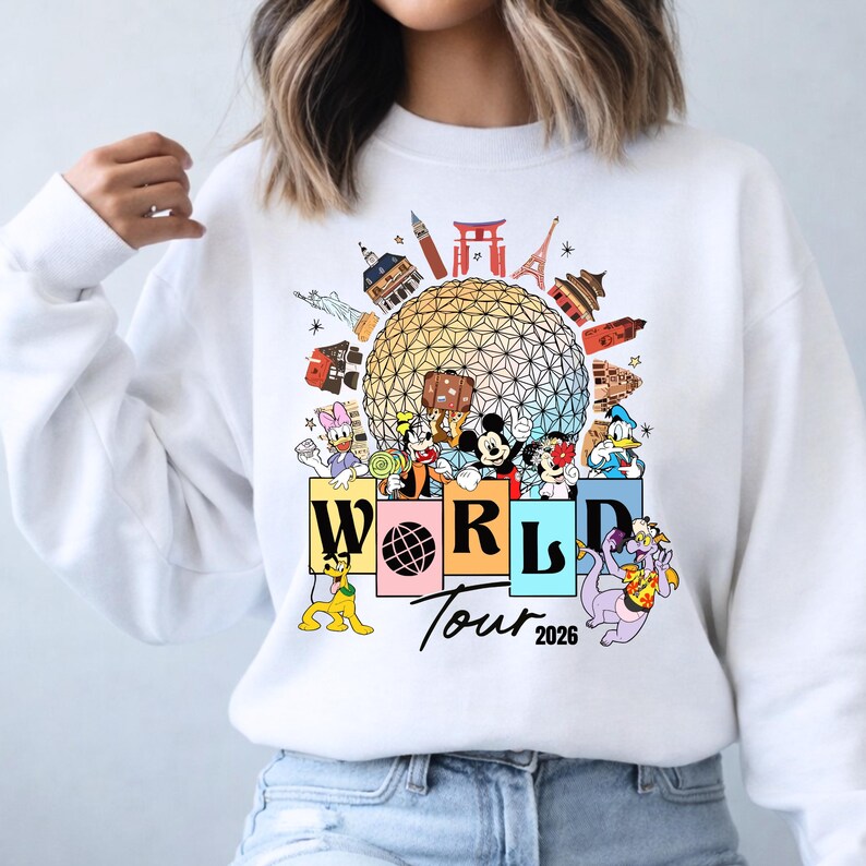 May include: White sweatshirt with a vibrant graphic of Disney characters and global landmarks. The design includes the text "WORLD Tour 2026", with Mickey Mouse, Donald Duck, and other characters. The design is set against a globe.