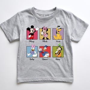 May include: A gray t-shirt featuring six Disney characters: Mickey Mouse, Minnie Mouse, Pluto, Daisy Duck, Donald Duck, and Goofy. Each character is in a colorful square with their name below. The shirt is made of a soft material.