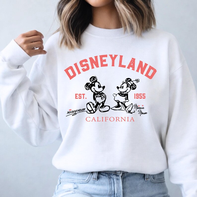 Puede incluir: Sudadera blanca con la palabra "DISNEYLAND" en letras rojas. Debajo de la palabra "DISNEYLAND" est&aacute;n las palabras "EST. 1955" y las palabras "Mickey Mouse" y "Minnie Mouse" en cursiva. La sudadera tambi&eacute;n tiene un gr&aacute;fico de Mickey y Minnie Mouse.
