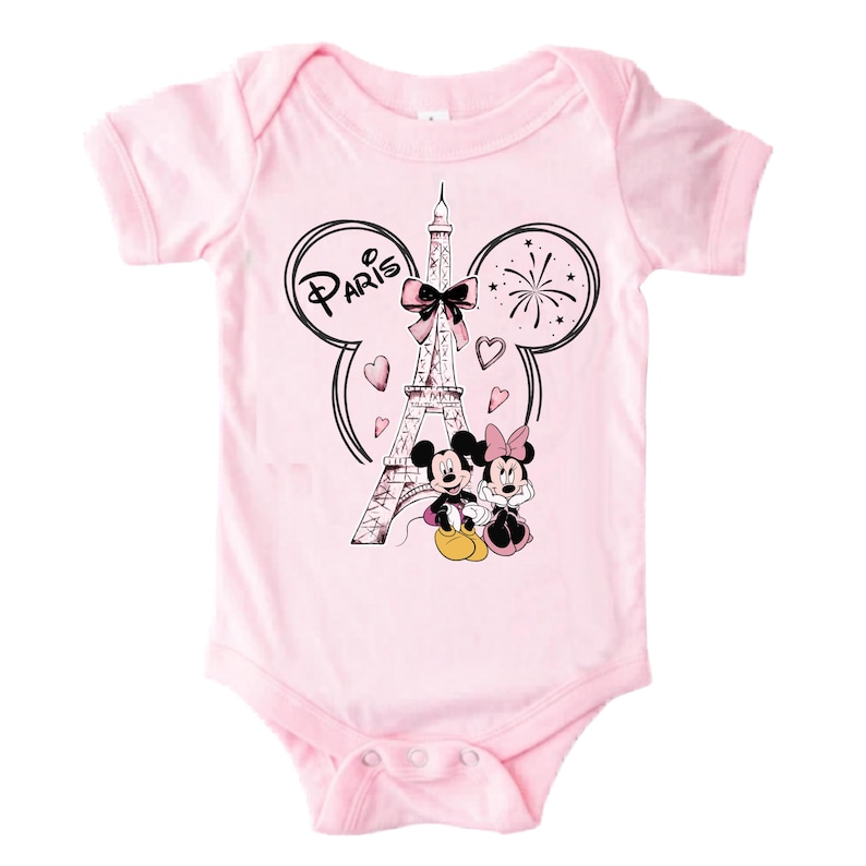 K&ouml;nnte beinhalten: Hellrosa Baby-Body mit einer Grafik von Mickey und Minnie Mouse vor dem Eiffelturm. Das Design enth&auml;lt das Wort "Paris" und Herzformen. Der Body hat kurze &Auml;rmel und einen runden Ausschnitt.