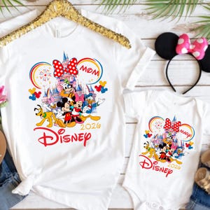 Op de afbeelding: Witte T-shirts en een babyromper met een Disney-ontwerp. Het ontwerp bevat Mickey en Minnie Mouse, Donald Duck en andere personages voor een kasteel. Het woord "Disney" is rood en het jaar "2020" is opgenomen.
