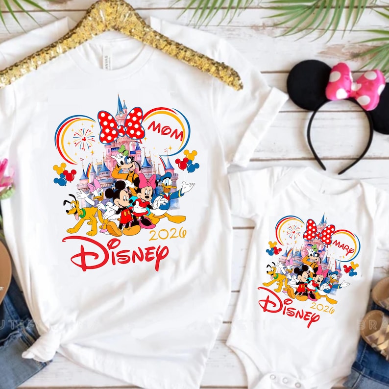 Op de afbeelding: Witte T-shirts en een babyromper met een Disney-ontwerp. Het ontwerp bevat Mickey en Minnie Mouse, Donald Duck en andere personages voor een kasteel. Het woord "Disney" is rood en het jaar "2020" is opgenomen.