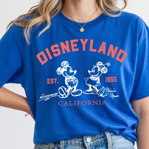 Puede incluir: Camiseta azul real con un gr&aacute;fico "DISNEYLAND" rojo desgastado. Presenta ilustraciones blancas de Mickey y Minnie Mouse, con "EST. 1955" y "CALIFORNIA" en rojo. Incluye los textos "Mickey Mouse" y "Minnie Mouse".