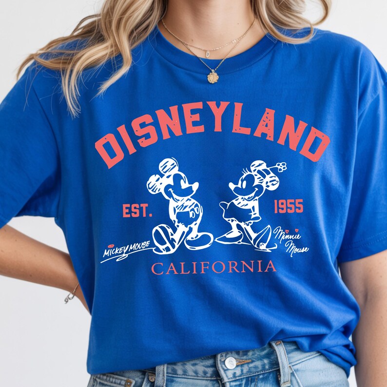 Puede incluir: Camiseta azul real con un gr&aacute;fico "DISNEYLAND" rojo desgastado. Presenta ilustraciones blancas de Mickey y Minnie Mouse, con "EST. 1955" y "CALIFORNIA" en rojo. Incluye los textos "Mickey Mouse" y "Minnie Mouse".