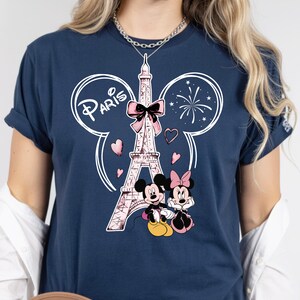 K&ouml;nnte beinhalten: Marineblaues T-Shirt mit einem rosa Eiffelturm-Motiv, Mickey und Minnie Mouse und dem Wort "Paris". Das Design beinhaltet Herzen und Feuerwerk. Das T-Shirt hat kurze &Auml;rmel.