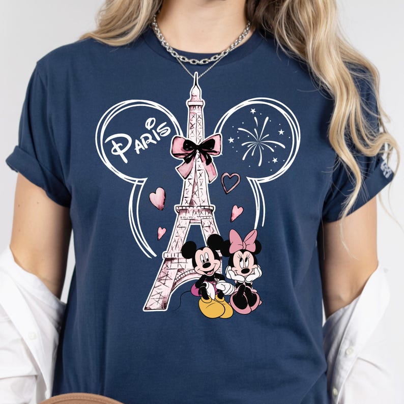 K&ouml;nnte beinhalten: Marineblaues T-Shirt mit einem rosa Eiffelturm-Motiv, Mickey und Minnie Mouse und dem Wort "Paris". Das Design beinhaltet Herzen und Feuerwerk. Das T-Shirt hat kurze &Auml;rmel.