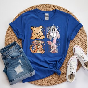 May include: Royal blue t-shirt featuring four cartoon character portraits: Winnie the Pooh, Eeyore, Tigger, and Piglet. The graphic is set within a grid of four squares. The shirt is paired with denim shorts and white canvas sneakers on a woven mat.