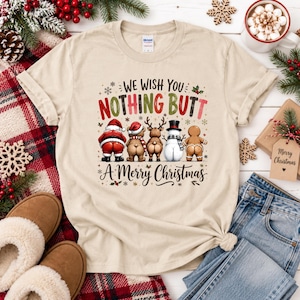 May include: A beige short-sleeve t-shirt featuring a graphic of five Christmas characters facing away with exposed buttocks. The text reads WE WISH YOU NOTHING BUTT A MERRY CHRISTMAS. The characters include Santa, a bear, a reindeer, a snowman, and a gingerbread man.