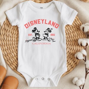 Puede incluir: Body de beb&eacute; blanco con un dise&ntilde;o de Disneyland en rojo y negro. El dise&ntilde;o presenta a Mickey y Minnie Mouse, el texto "Disneyland", "Est. 1955" y "California".