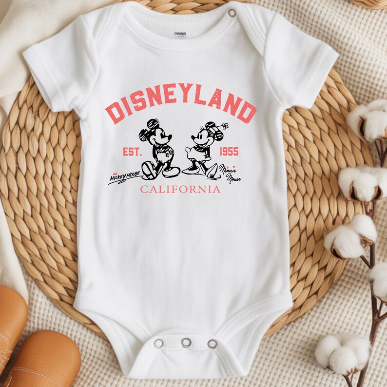 Puede incluir: Body de beb&eacute; blanco con un dise&ntilde;o de Disneyland en rojo y negro. El dise&ntilde;o presenta a Mickey y Minnie Mouse, el texto "Disneyland", "Est. 1955" y "California".