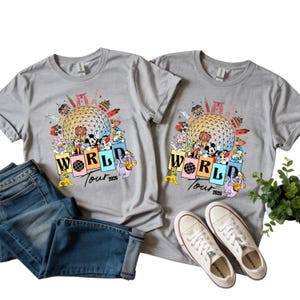 May include: Two grey t-shirts with a colourful graphic featuring Disney characters, a globe, and the text "WORLD Tour 2026." The shirts are paired with blue jeans and white canvas trainers.