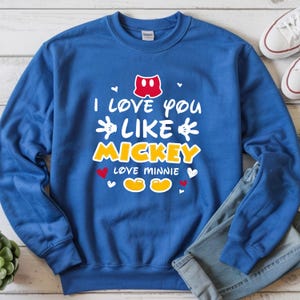 Pode incluir: Camisola azul com o texto "I love you like Mickey, love Minnie" em branco e amarelo. O design inclui cal&ccedil;&otilde;es do Mickey Mouse, cora&ccedil;&otilde;es e os sapatos do Mickey e da Minnie. Cal&ccedil;as de ganga e t&eacute;nis brancos s&atilde;o vis&iacute;veis.