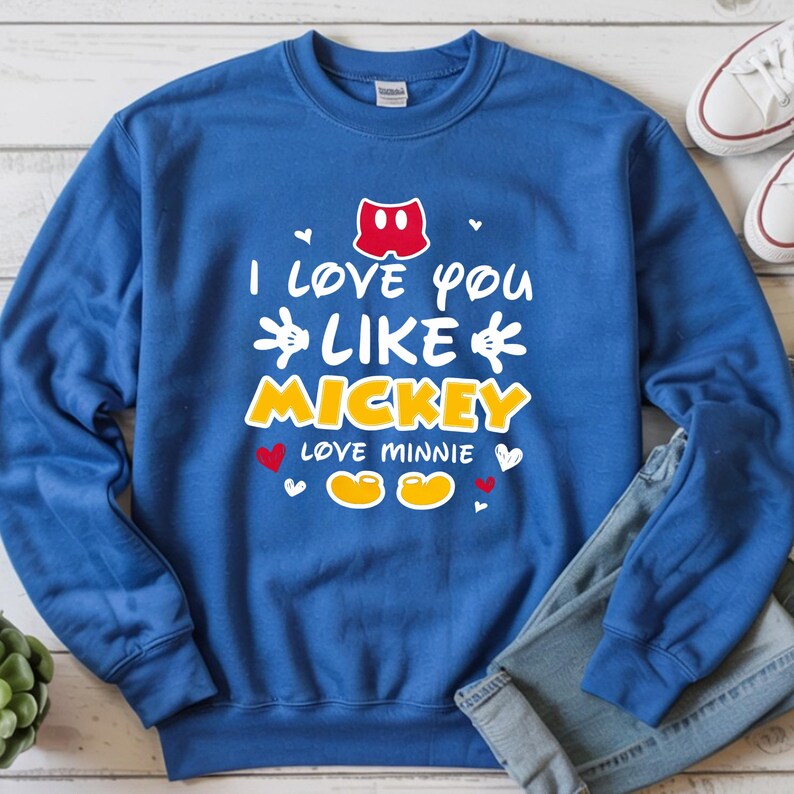 Pode incluir: Camisola azul com o texto "I love you like Mickey, love Minnie" em branco e amarelo. O design inclui cal&ccedil;&otilde;es do Mickey Mouse, cora&ccedil;&otilde;es e os sapatos do Mickey e da Minnie. Cal&ccedil;as de ganga e t&eacute;nis brancos s&atilde;o vis&iacute;veis.