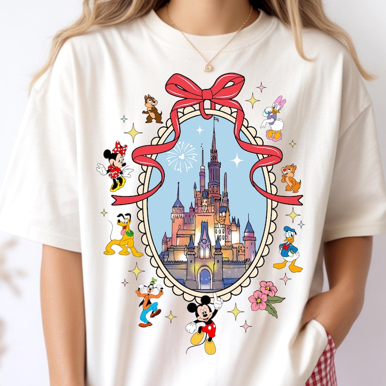 May include: Cream-colored t-shirt featuring a colorful graphic of a castle inside a decorative frame, surrounded by cartoon characters like Mickey Mouse, Minnie Mouse, Donald Duck, and Pluto. A red ribbon tops the frame. The design includes stars and flowers.
