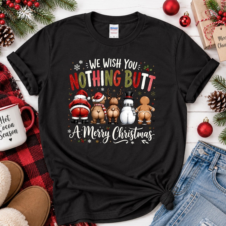May include: A black short-sleeve t-shirt featuring a graphic of five Christmas characters facing away, with the text "WE WISH YOU NOTHING BUTT A MERRY CHRISTMAS" in red, green, and white lettering, surrounded by small white snowflake accents.