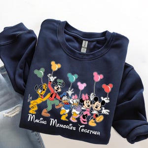 May include: Navy blue sweatshirt featuring a graphic of Disney characters Pluto, Goofy, Donald Duck, Daisy Duck, Minnie Mouse, and Mickey Mouse holding Mickey Mouse-shaped balloons. The text "Making Memories Together" is printed below the characters.