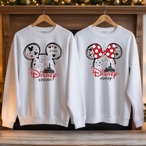 May include: Two white sweatshirts with Disney designs. One has Mickey Mouse ears, fireworks, and the name "Steven"; the other has Minnie Mouse ears, a red polka-dot bow, and the name "Ashley." Both include the year 2020.