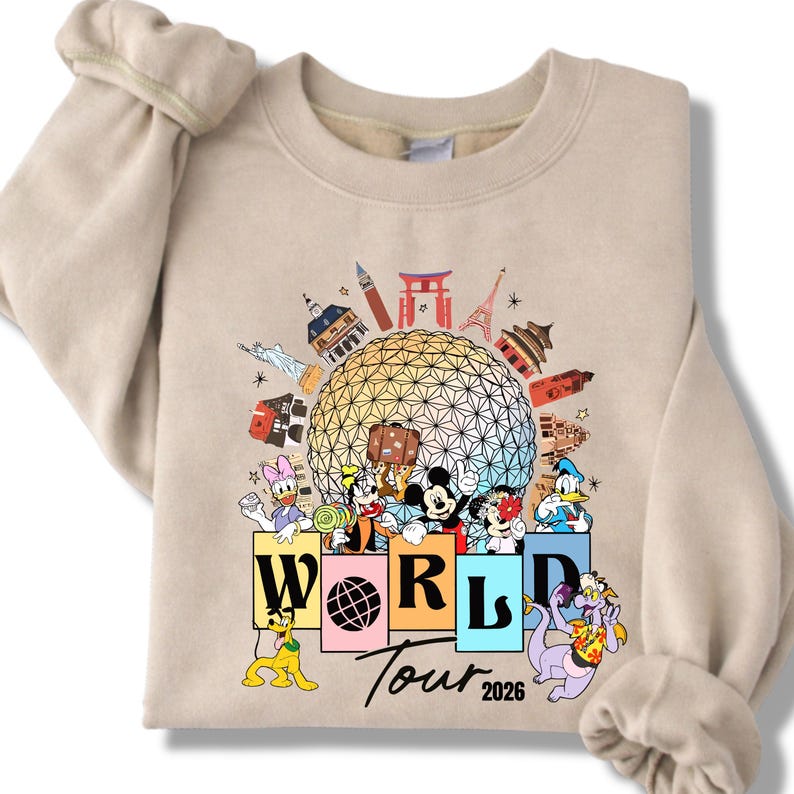 May include: Beige sweatshirt with a colourful graphic of Disney characters and landmarks. The design includes Mickey Mouse, Donald Duck, and other characters, with the text "WORLD Tour 2026". The sweatshirt has long sleeves.
