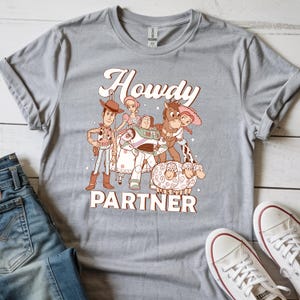 May include: Gray t-shirt featuring a graphic of characters from a popular animated film. The design includes the text "Howdy Partner" in a vintage font. The shirt is paired with blue jeans and white sneakers.