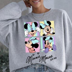 Pu&ograve; includere: Una felpa grigia con un design a quattro pannelli di Minnie Mouse in varie pose, ognuna con un fiocco azzurro. Il design include il testo corsivo "Minnie Mouse" con "xoxo" sotto.