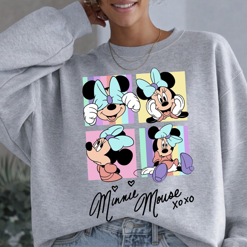 Pu&ograve; includere: Una felpa grigia con un design a quattro pannelli di Minnie Mouse in varie pose, ognuna con un fiocco azzurro. Il design include il testo corsivo "Minnie Mouse" con "xoxo" sotto.