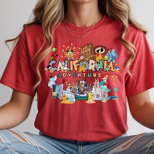 May include: Red t-shirt featuring the words "CALIFORNIA ADVENTURE" in a colourful graphic, with cartoon characters and amusement park imagery. The design includes a Ferris wheel, roller coaster, and castle.