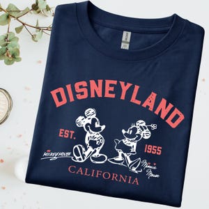 Puede incluir: Camiseta azul marino con la palabra "DISNEYLAND" en letras rojas. Debajo, ilustraciones blancas de Mickey y Minnie Mouse, con los textos "EST. 1955" y "CALIFORNIA". La camiseta es de la marca Gildan.