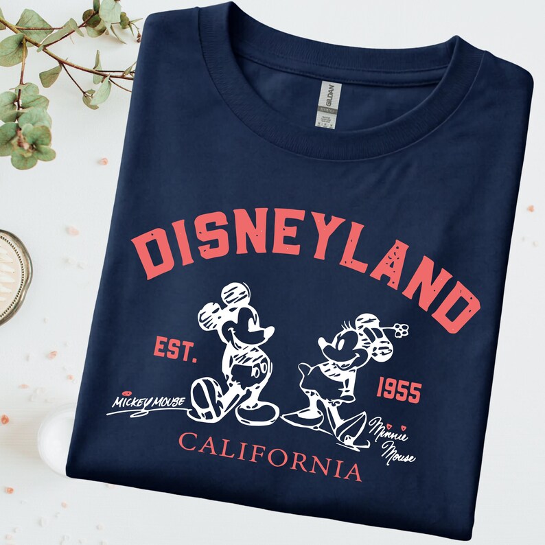 Puede incluir: Camiseta azul marino con la palabra "DISNEYLAND" en letras rojas. Debajo, ilustraciones blancas de Mickey y Minnie Mouse, con los textos "EST. 1955" y "CALIFORNIA". La camiseta es de la marca Gildan.