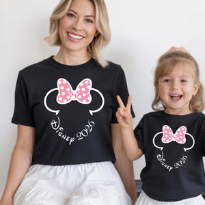 May include: Black t-shirts featuring a Minnie Mouse design with a pink polka dot bow, white outline, and the text "Disney 2026". The shirts are worn by a woman and a child.