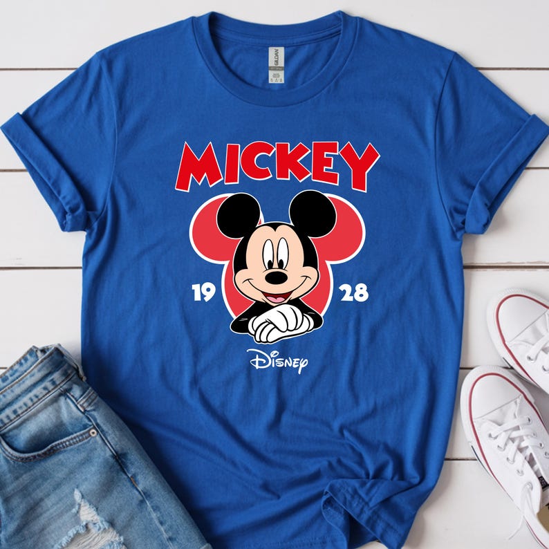 May include: A royal blue t-shirt featuring a Mickey Mouse graphic. The design includes the word "MICKEY" in red, the character's face, and the years "19" and "28". The Disney logo is at the bottom. White sneakers and blue jeans are visible.