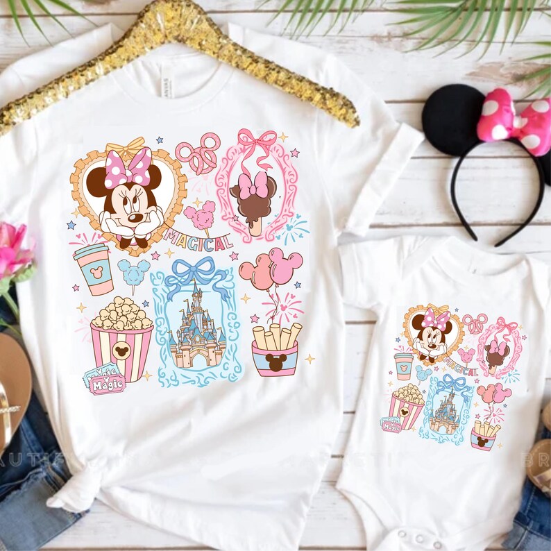 Puede incluir: Camisetas blancas con un dise&ntilde;o de dibujos animados colorido con Minnie Mouse, palomitas de ma&iacute;z, un castillo y la palabra "Magical". Una camiseta es para adultos y la otra es un body para beb&eacute;. Tambi&eacute;n se ve una diadema con orejas de Minnie Mouse con un lazo rosa.
