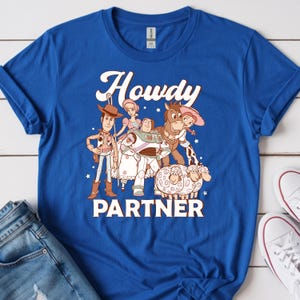 May include: Royal blue t-shirt featuring a graphic of characters from a popular animated movie. The word "Howdy" is above the characters, and the word "Partner" is below. The shirt is paired with blue jeans and white sneakers.