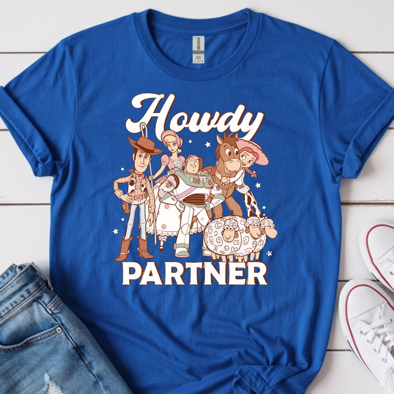 May include: Royal blue t-shirt featuring a graphic of characters from a popular animated movie. The word "Howdy" is above the characters, and the word "Partner" is below. The shirt is paired with blue jeans and white sneakers.