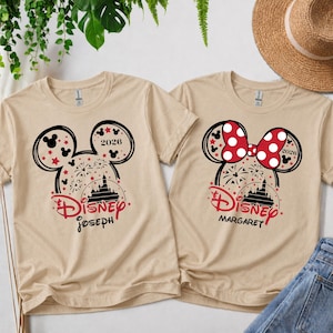 May include: Two beige t-shirts with Disney designs. One displays a Mickey Mouse head with fireworks, stars, and the name "Joseph". The other has a Minnie Mouse bow and the name "Margaret". Both shirts include the year "2026".