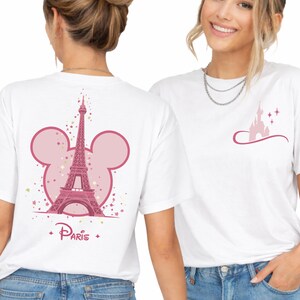 May include: White t-shirts with pink graphic designs. One shirt features the Eiffel Tower inside a Mickey Mouse head outline with the word "Paris". The other shirt has a pink castle design with stars.